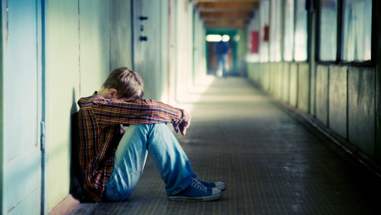 Teenager curled up in a ball leaning against lockers in a school with their head in their arms.