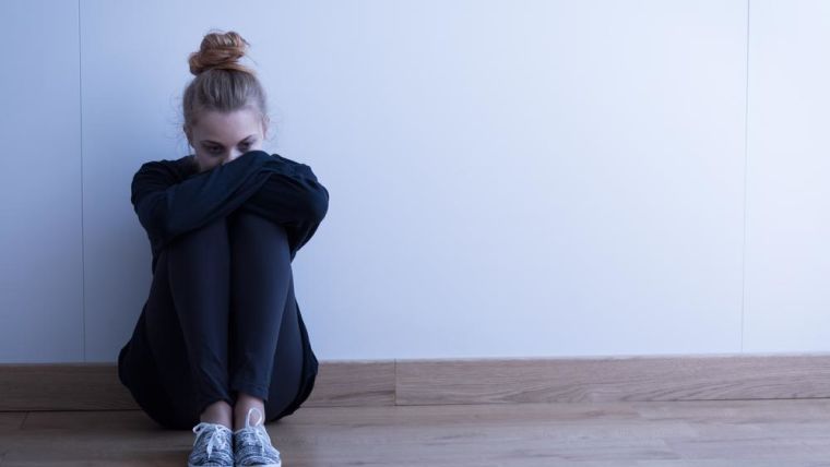 Image shows girl sat with her head on her knees against the wall looking sad.