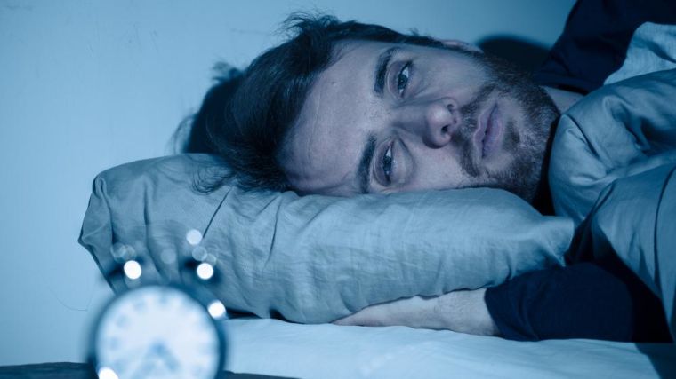 A man with tired eyes laying down in bed with an alarm clock blurred in the image.