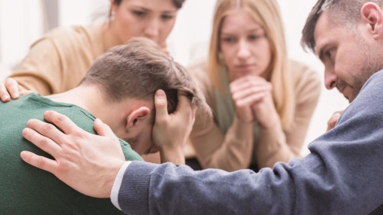 Close-up of a devastated young man holding his head in his hands and friends supporting him during group therapy