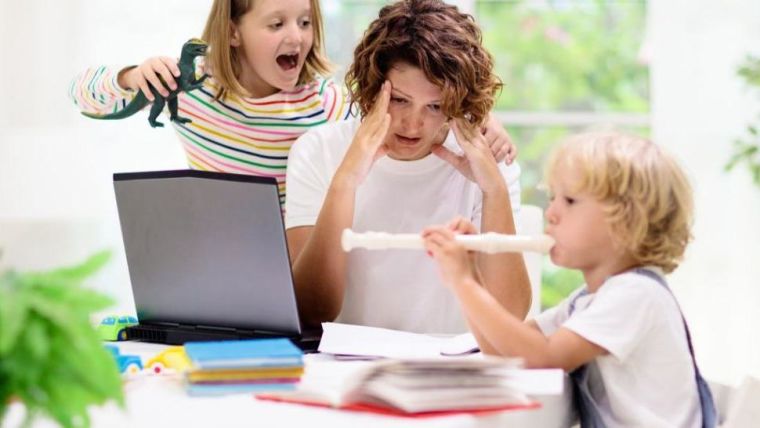 Image shows women stressed because she is trying to work whilst her young children are distracting her with noise and youthful energy.