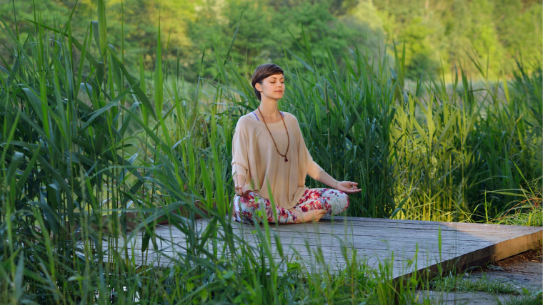 Image shows woman sat in a meditative posture surrounded by green vegetation.
