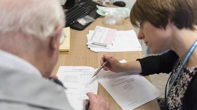 Image shows a medical staff helping an elderly man fill out paperwork.