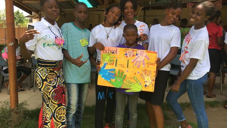 Image shows group of African people, some adults, children and teenagers, smiling and holding up a colourful piece of artwork one of the children has created.