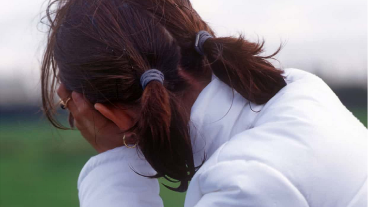 Image shows a young girl facing away from the camera with her head in her hands.