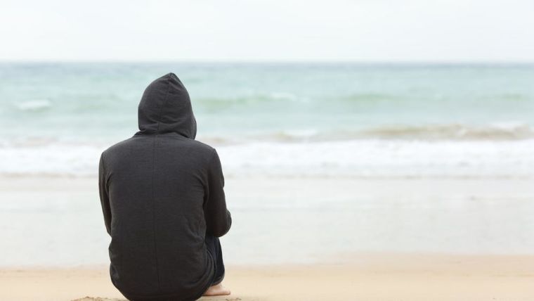 A boy in a hoodie sitting on a beach looking out to sea