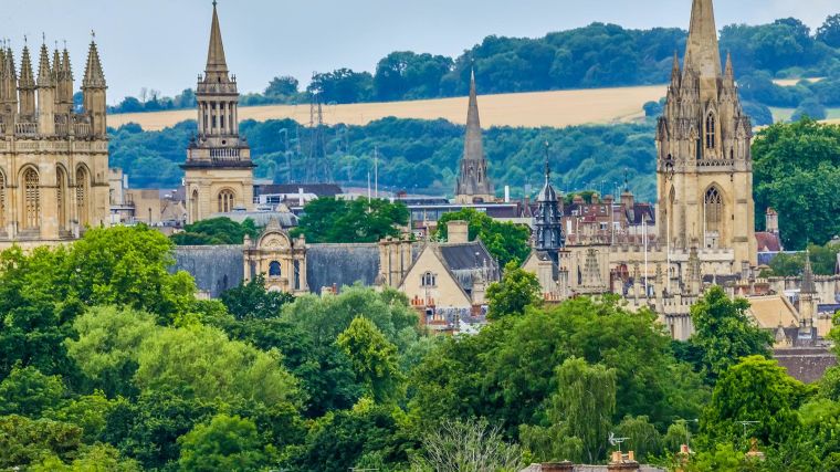 Image of Oxford city spires skyline including some tree-line and hills in the backdrop.