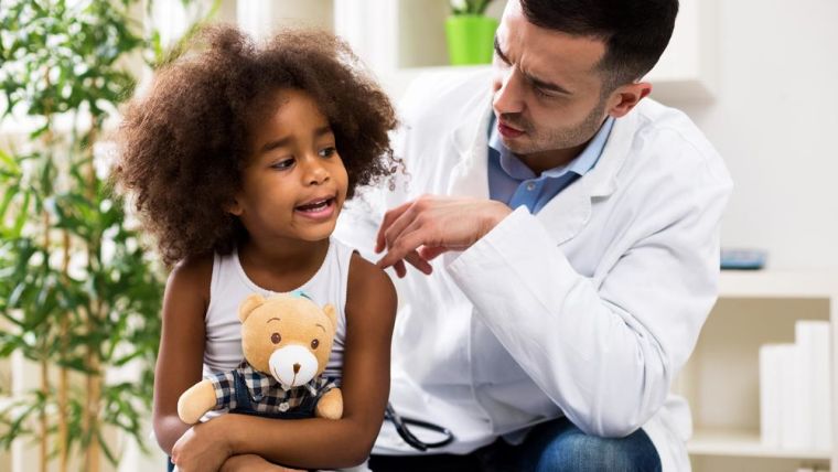 Image shows young child holding a teddie bear talking to a doctor who is knelt beside her.
